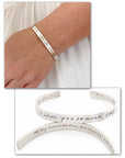 Silver bracelet with engraved text on a wrist, displayed with additional bracelets on a white background.