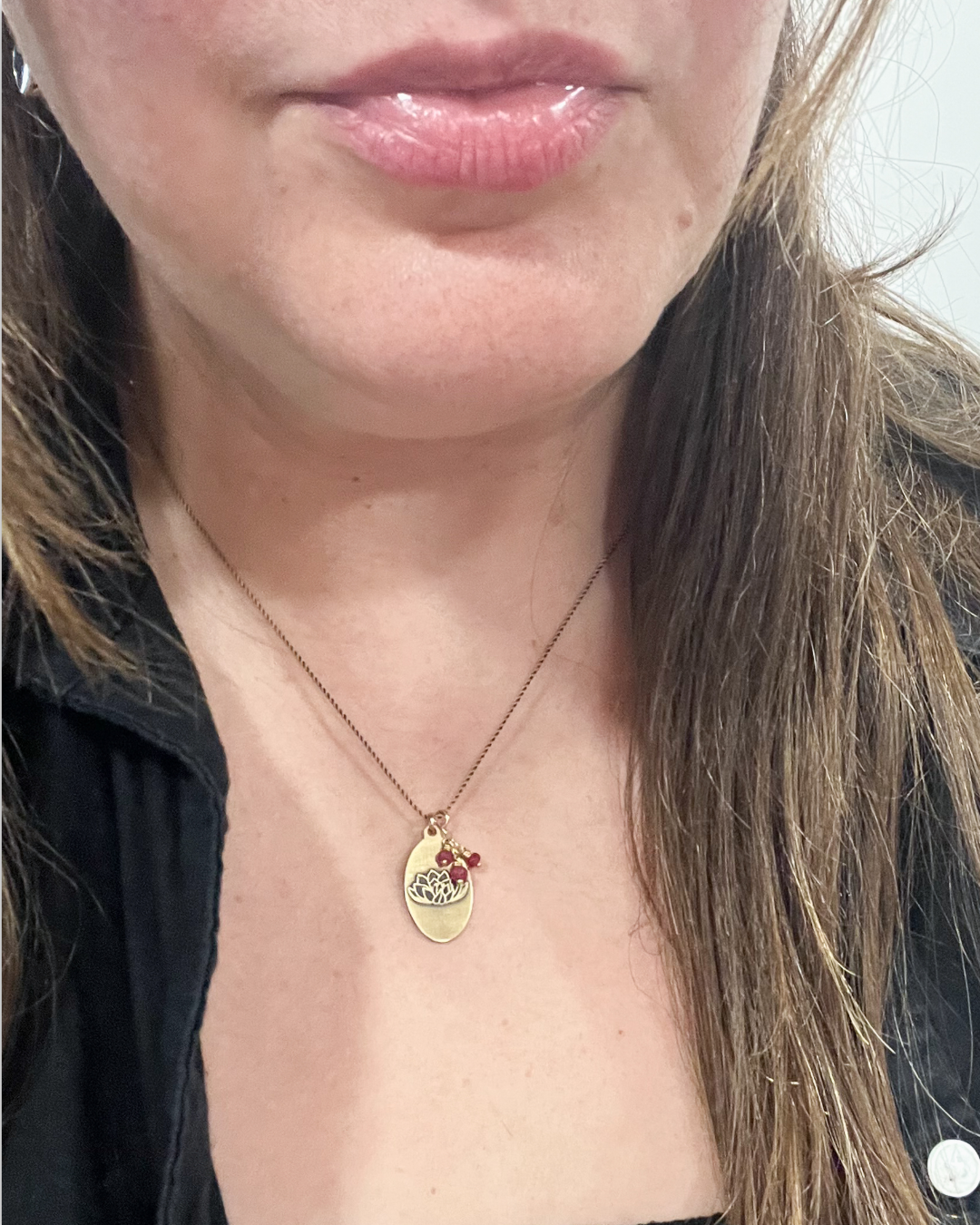 A close-up of a person wearing a delicate gold necklace with a small oval pendant and tiny red and white details, shown against the neck and upper chest.