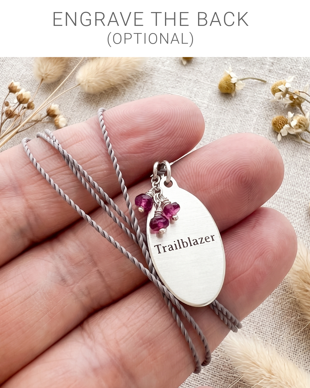 A close-up of a hand holding a silver oval pendant necklace engraved with the word 'Trailblazer,' attached to a gray braided cord, with small pink gemstone charms. Text at the top reads 'ENGRAVE THE BACK (OPTIONAL),' and dried flowers are scattered in the background on a light fabric surface.