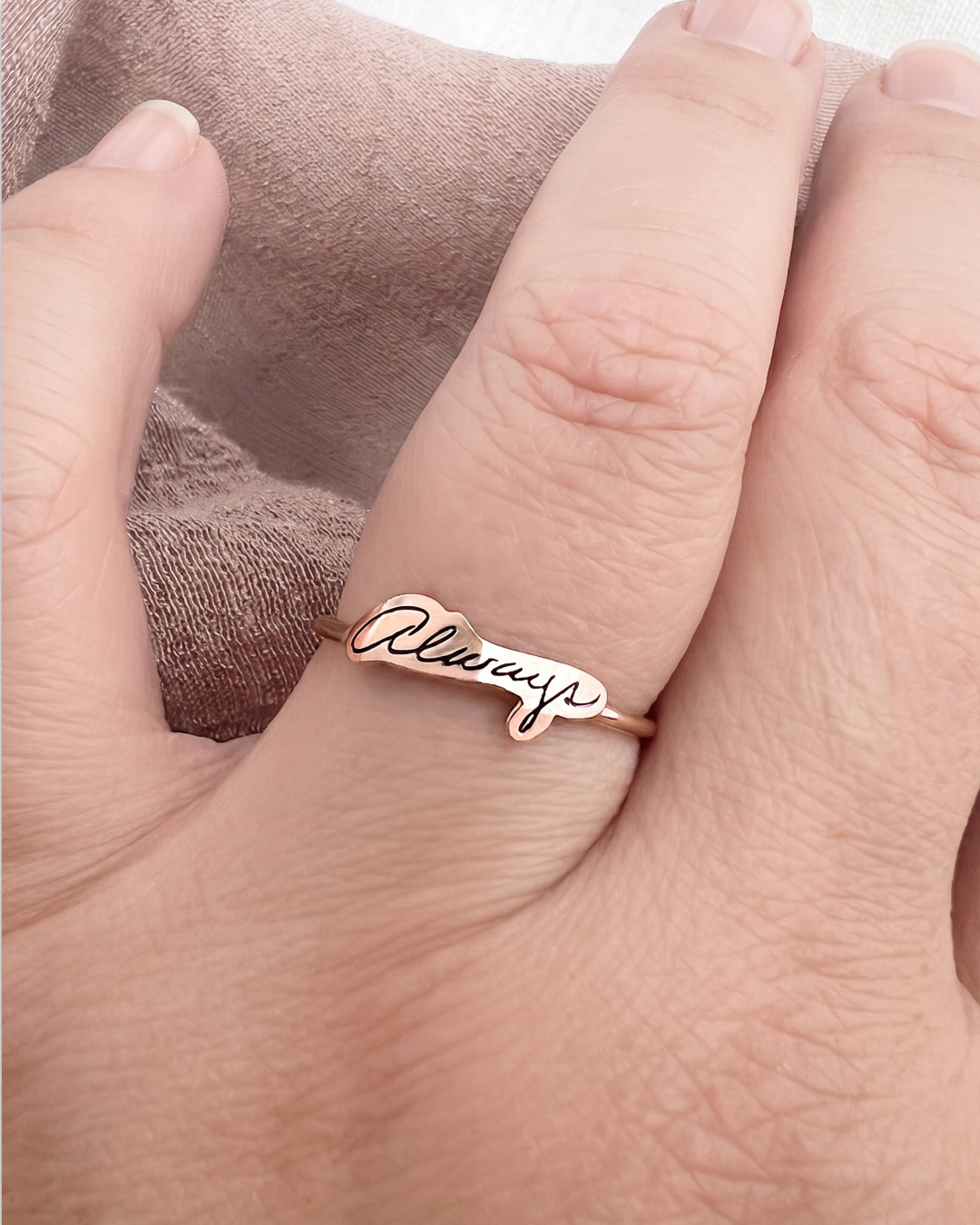 A close-up of a hand wearing a rose gold script name ring that reads 'Always'.