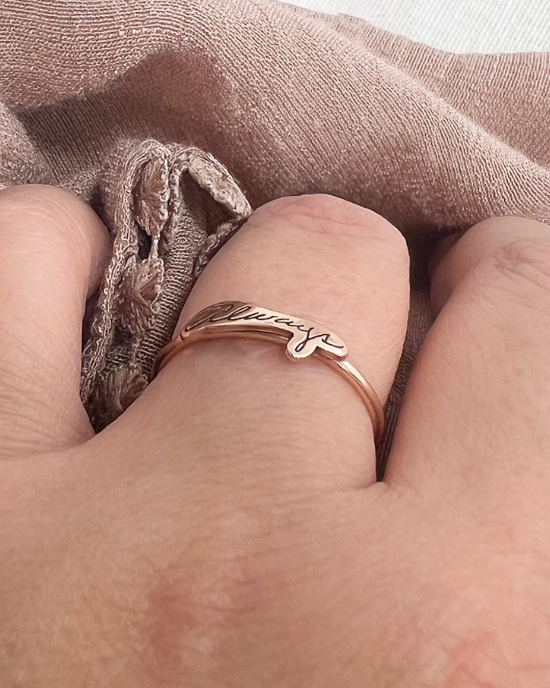 A close-up of a hand wearing a bronze band ring with the word 'Always' engraved on it, resting against a dusty rose fabric background.
