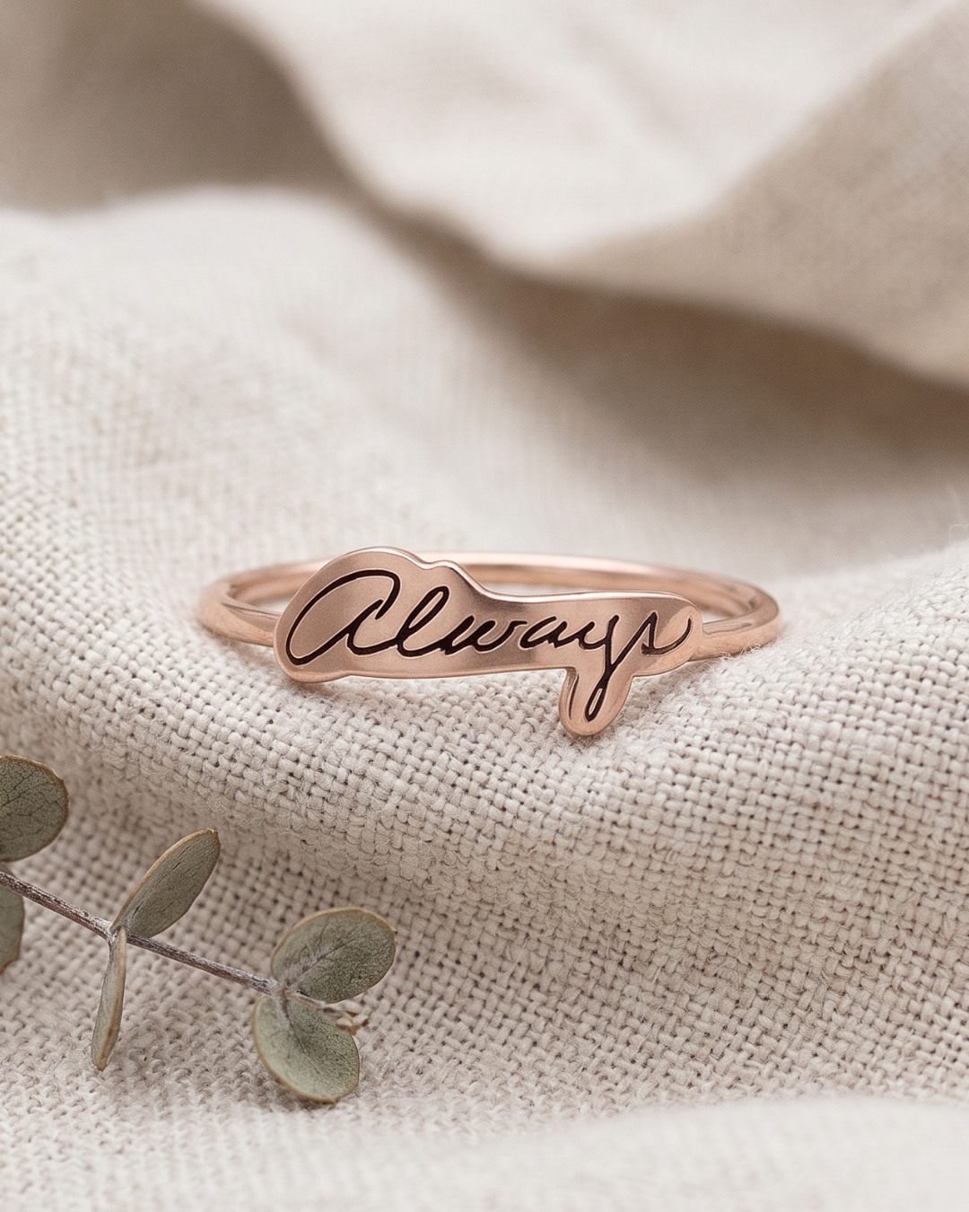 A bronze open band ring engraved with the word 'Always' in black handwritten script, resting on a textured beige linen fabric with a sprig of eucalyptus in the foreground.