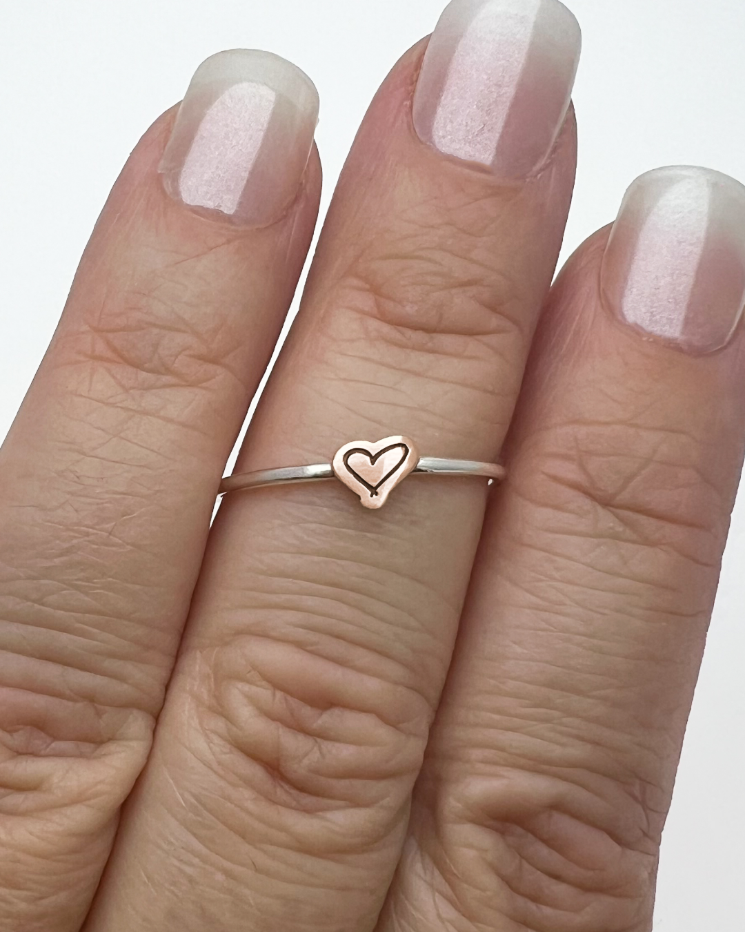 A close-up of a hand wearing a delicate rose gold ring with a small heart outline charm, set against a clean white background.
