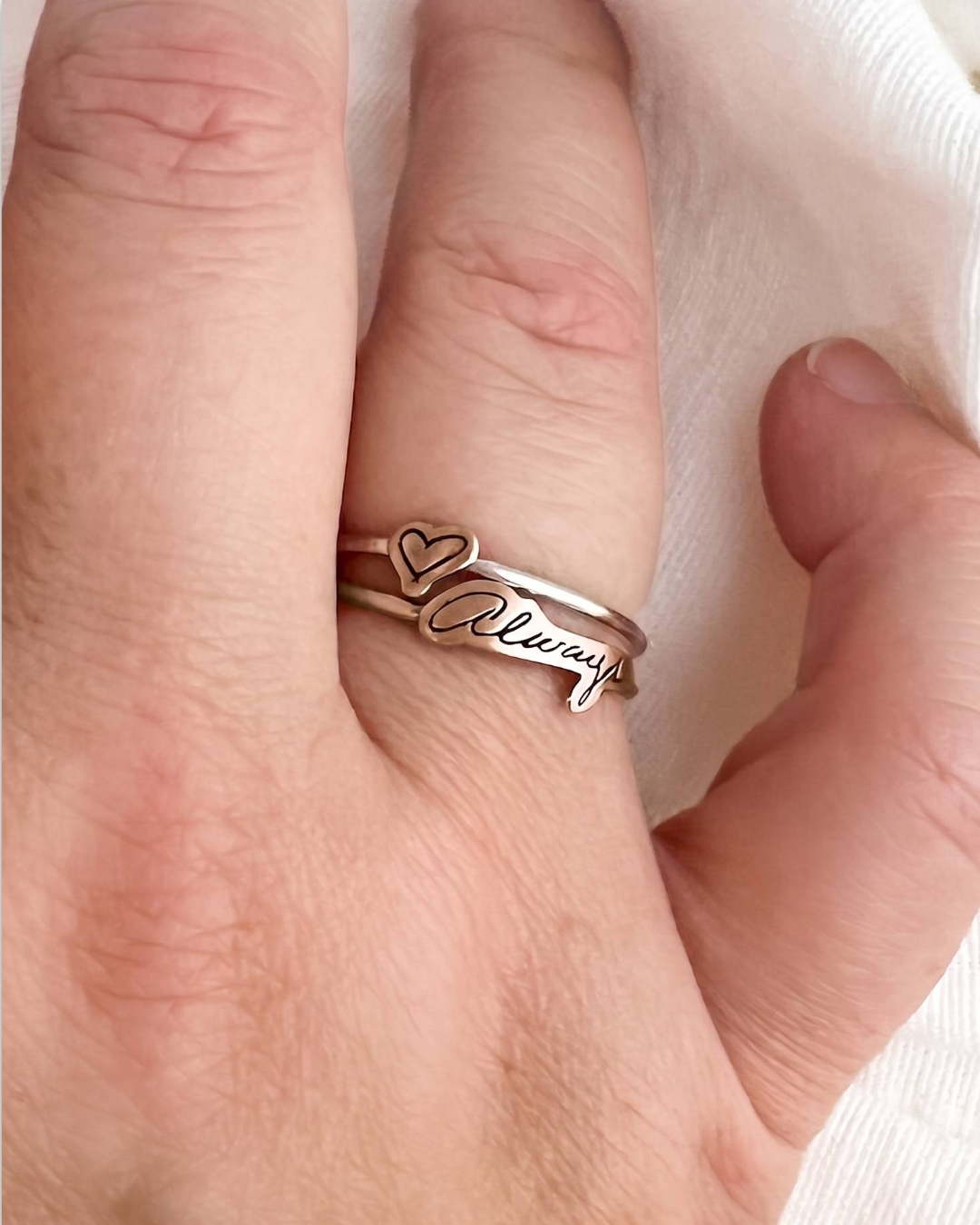 A close-up of a hand wearing a double-band silver ring with a heart motif and the word 'Always' engraved on the front, resting on a soft white fabric background.