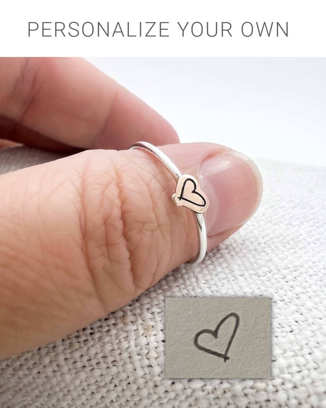 A close-up of a hand wearing a thin silver ring with a small heart-shaped charm outlined in bronze The image includes a text overlay at the top reading 'PERSONALIZE YOUR OWN' and a small inset showing a heart design stamped on a metal surface.
