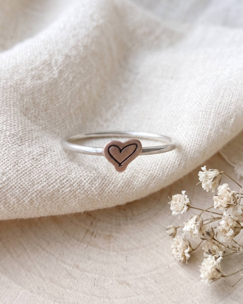 A thin silver band ring with a small heart-shaped charm featuring a double-lined heart design, resting on a textured cream linen fabric next to dried white flowers.