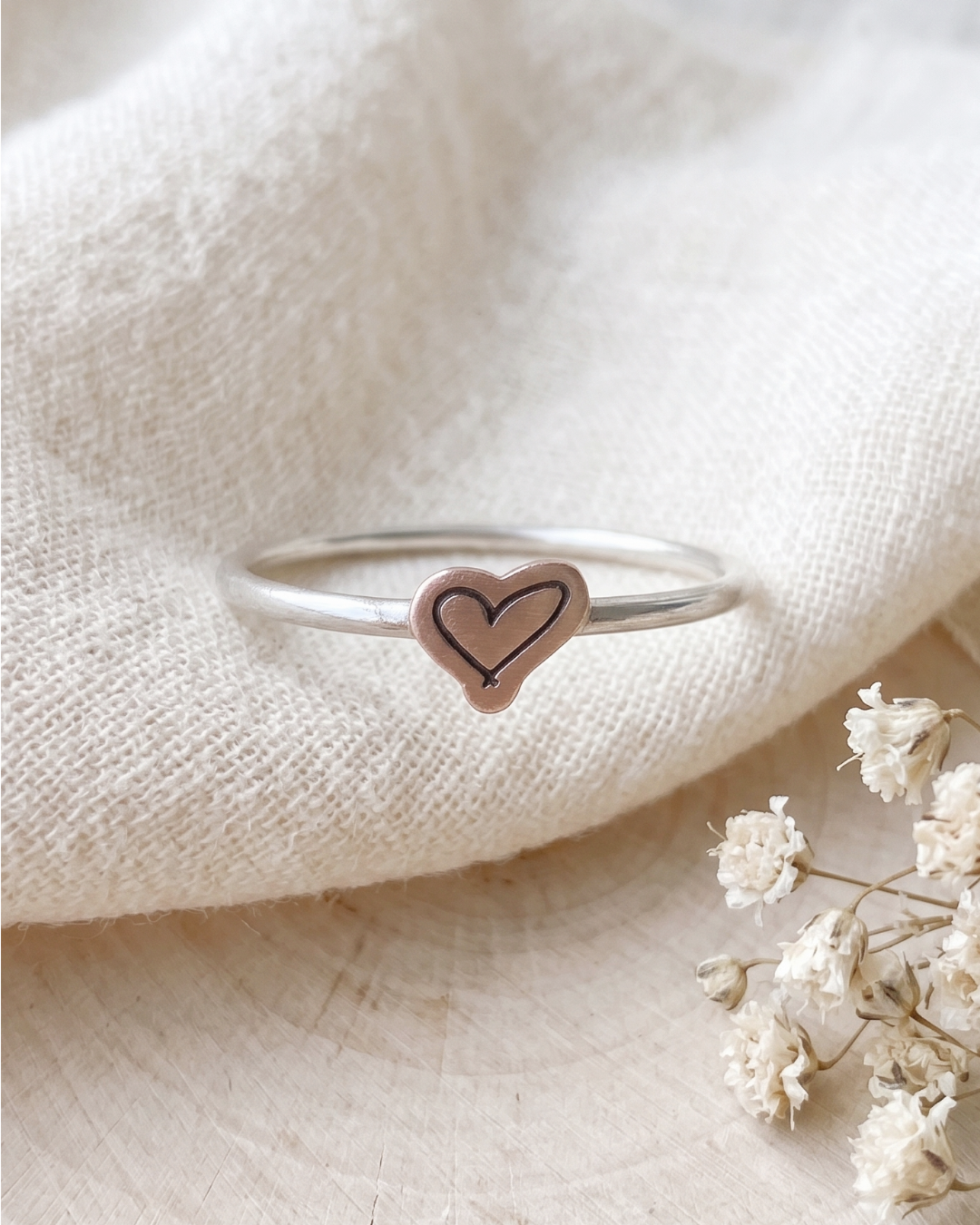 A silver-toned ring with a small heart-shaped charm featuring a black hand-drawn line design, resting on a textured cream fabric with dried white flowers in the corner.