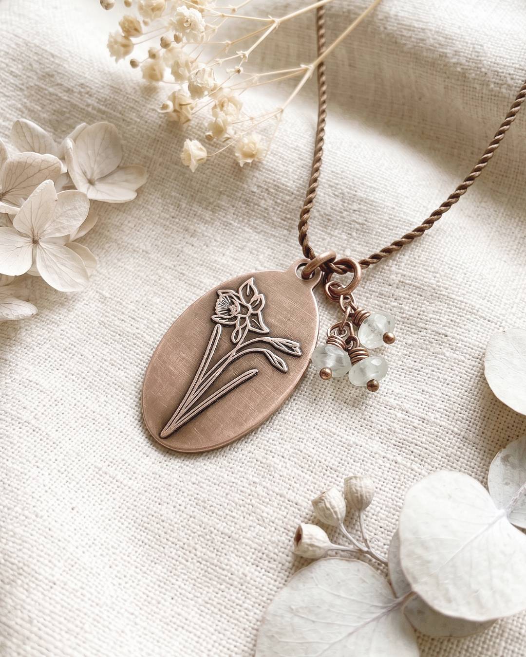 An oval bronze pendant necklace with a stamped flower design, hanging from a twisted brown cord with small clear bead charms, styled on a textured beige linen fabric with dried flowers.