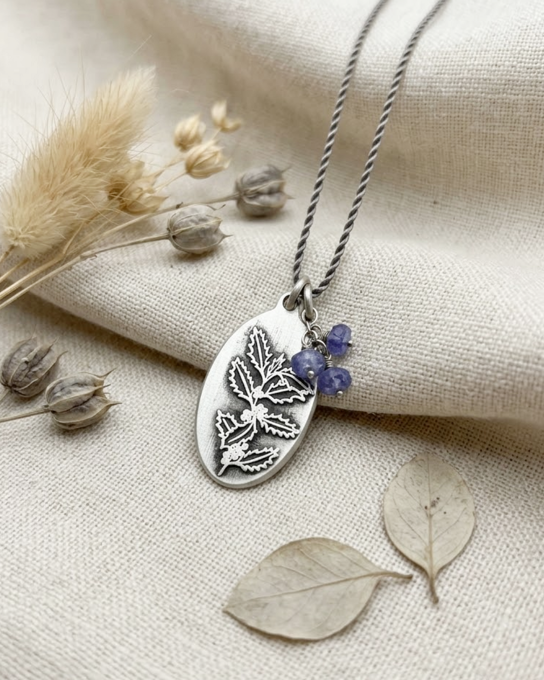 A silver oval pendant necklace with an engraved holly design and tanzanite bead accents, displayed on a textured beige fabric with dried botanical props.