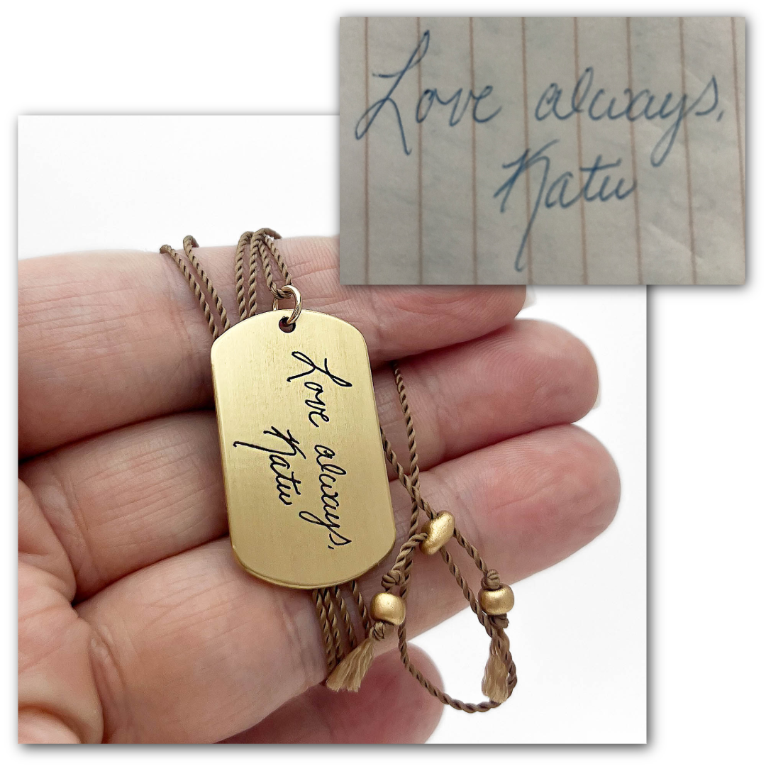 Gold toned brass necklace with engraved message held by a hand, with a handwritten note in the background.
