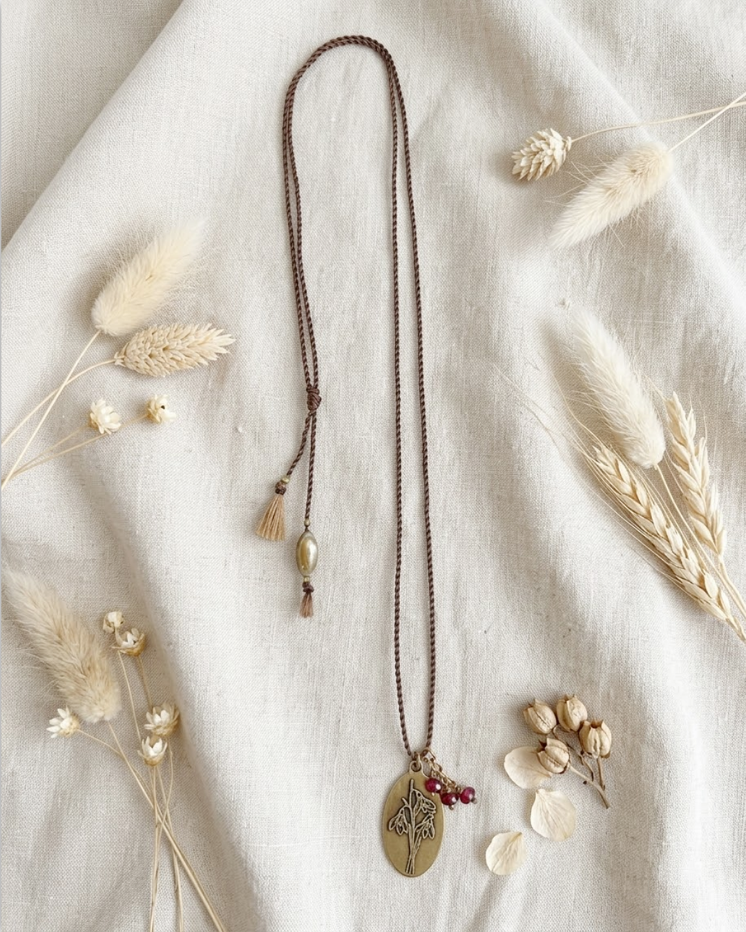 A bohemian-style long necklace with a dark brown beaded chain, a round gold-toned pendant featuring a snowdrop flower design, and small red bead accents, styled on a cream linen fabric with dried grasses.