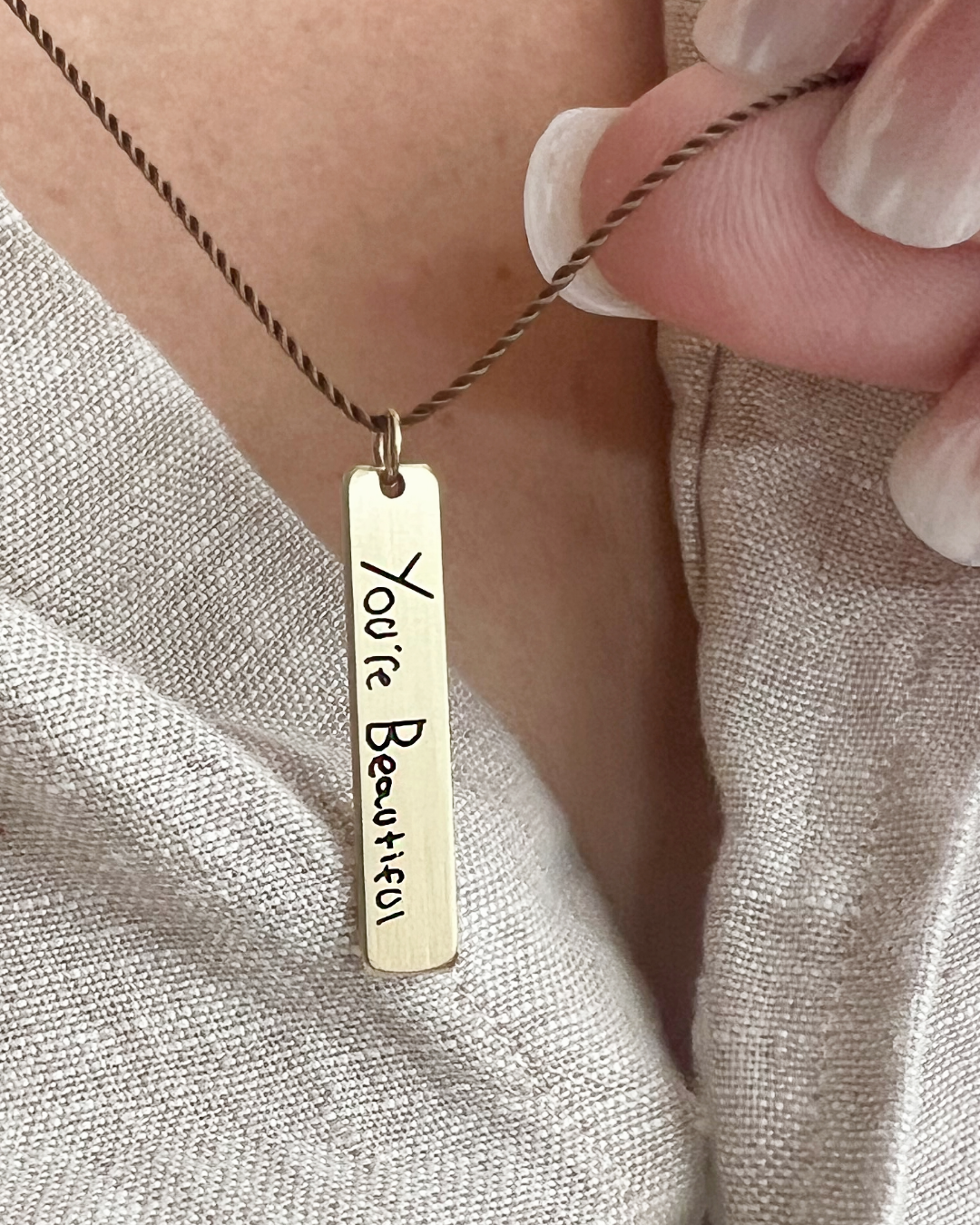 Gold memorial necklace with a pendant reading 'You're Beautiful' in handwriting, held by a hand against a neutral background.