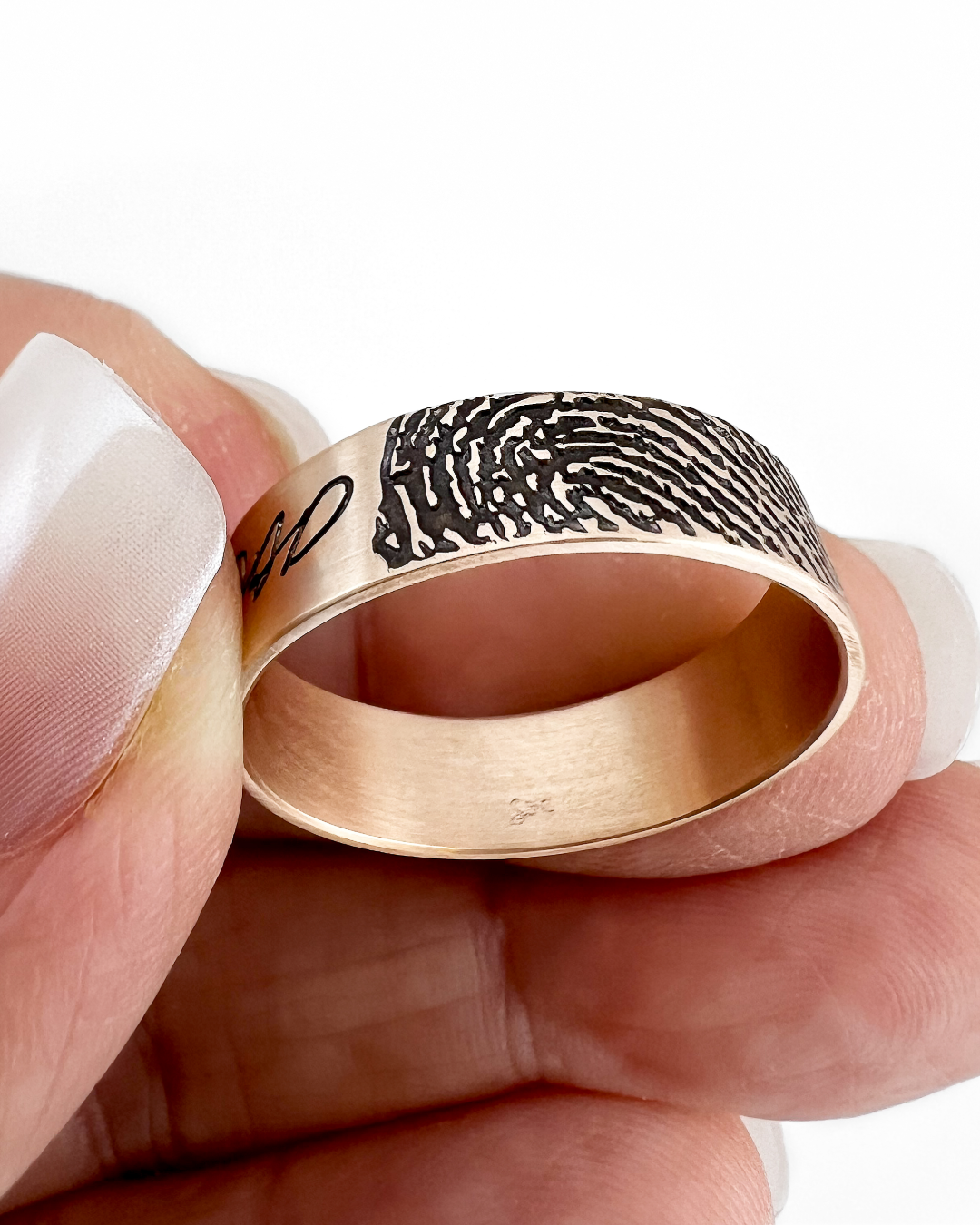 A close-up of a hand holding a custom engraved ring. The ring has a brushed rose gold or copper-toned metal band with a black oxidized fingerprint pattern on one side and a black script engraving on the other.