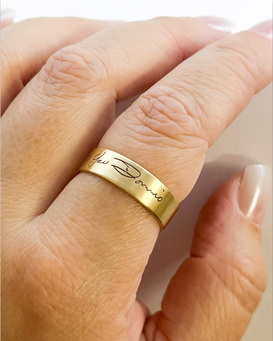A close-up of a hand wearing a polished gold ring engraved with the cursive script.