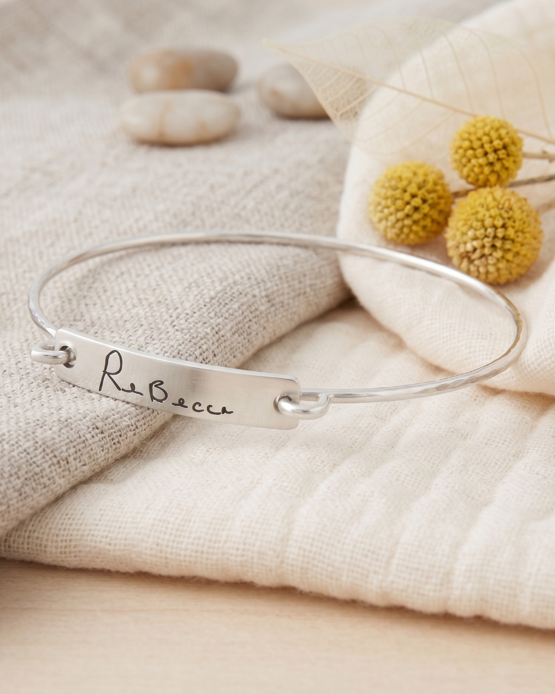 A silver bangle bracelet with a custom engraved name plate reading 'Rebecca', styled on a textured cream fabric with dried yellow flowers and neutral stones in the background.