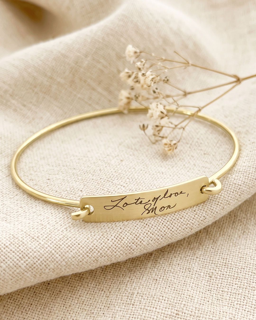 A brass bangle bracelet with a rectangular engraved nameplate reading 'Lots of Love, Mom' resting on a textured beige linen cloth with dried baby's breath flowers.