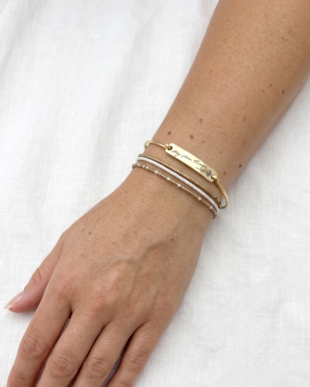 A close-up of a person's wrist wearing three delicate bracelets against a soft white fabric background. The bracelets include a gold-tone engraved bangle, a thin white bangle, and a textured gold bangle.