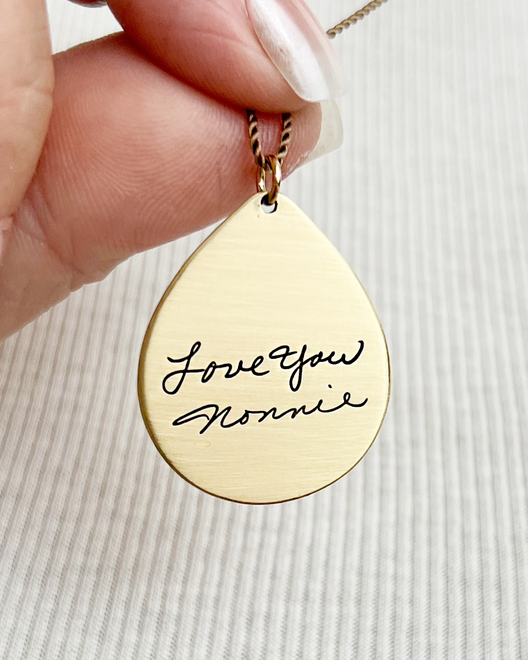 Brass teardrop pendant with 'Love you Nonnie' engraving of clients handwriting held by a hand.