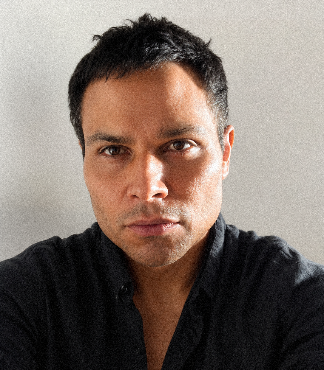 A close-up portrait of a man with short dark hair wearing a dark button-up shirt against a plain light gray background.