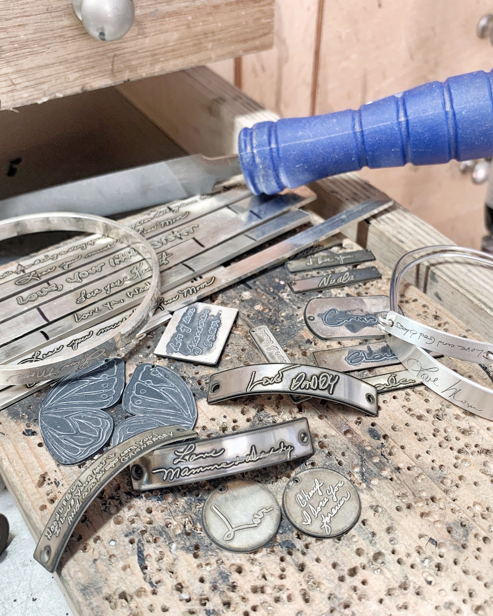 A collection of custom-engraved silver metal bracelets and nameplates displayed on a workbench next to a blue-handled tool.