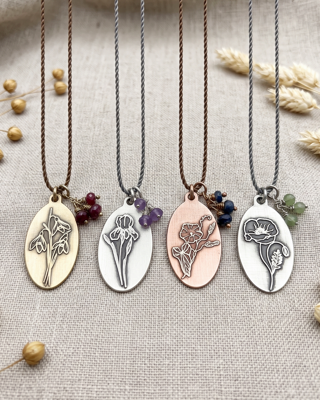 Four oval pendant necklaces with floral engravings and small bead charms, displayed on a textured beige fabric background with dried flowers. The necklaces have gold, silver, rose gold, and white metal pendants with brown, purple, blue, and green beads.