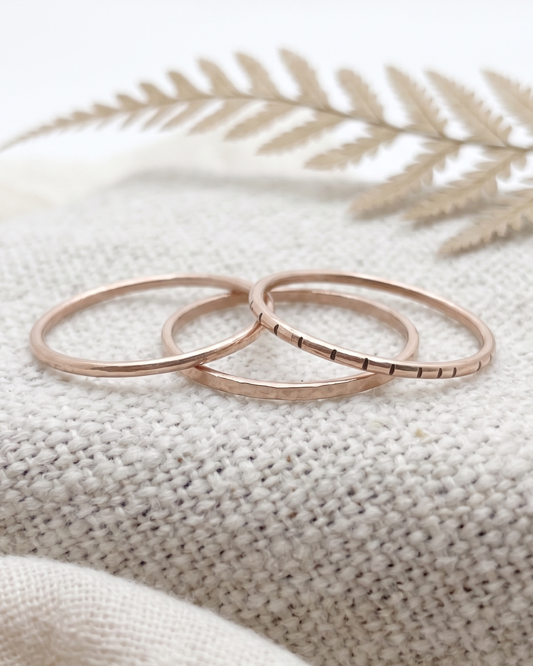 Three thin bronze rings with textured, ridged bands arranged on a beige woven fabric with a dried fern leaf in the background.