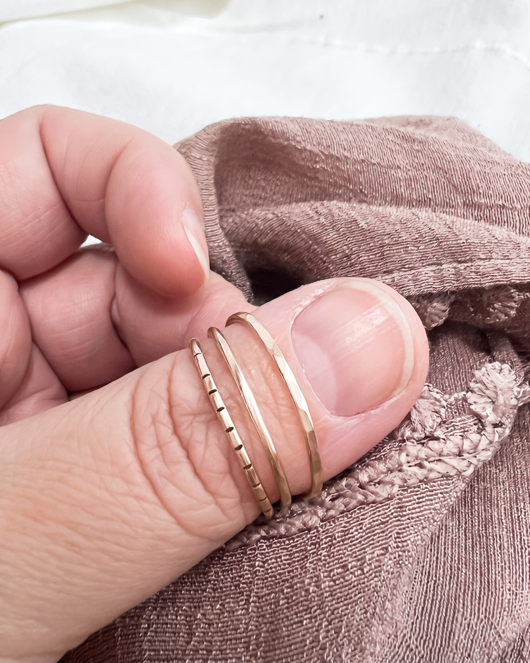 Three delicate rose gold colored rings worn on a person's thumb, shown close-up against a soft brown fabric background.