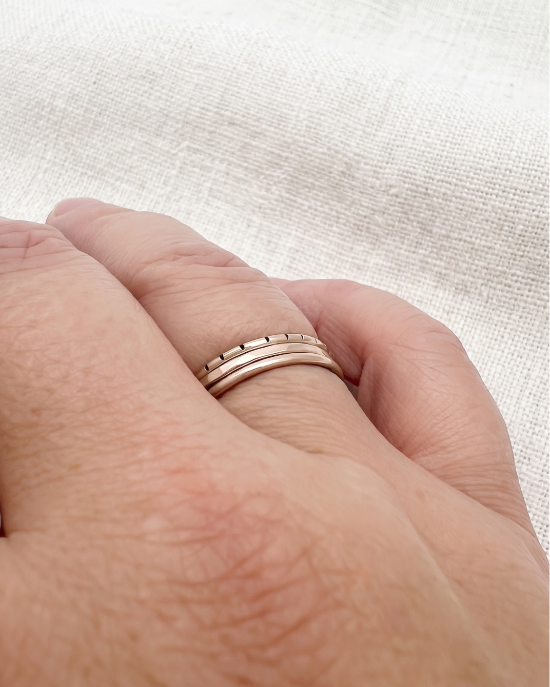 A close-up of a person's hand wearing three slim rose gold stacking rings, set against a neutral, textured beige linen background.