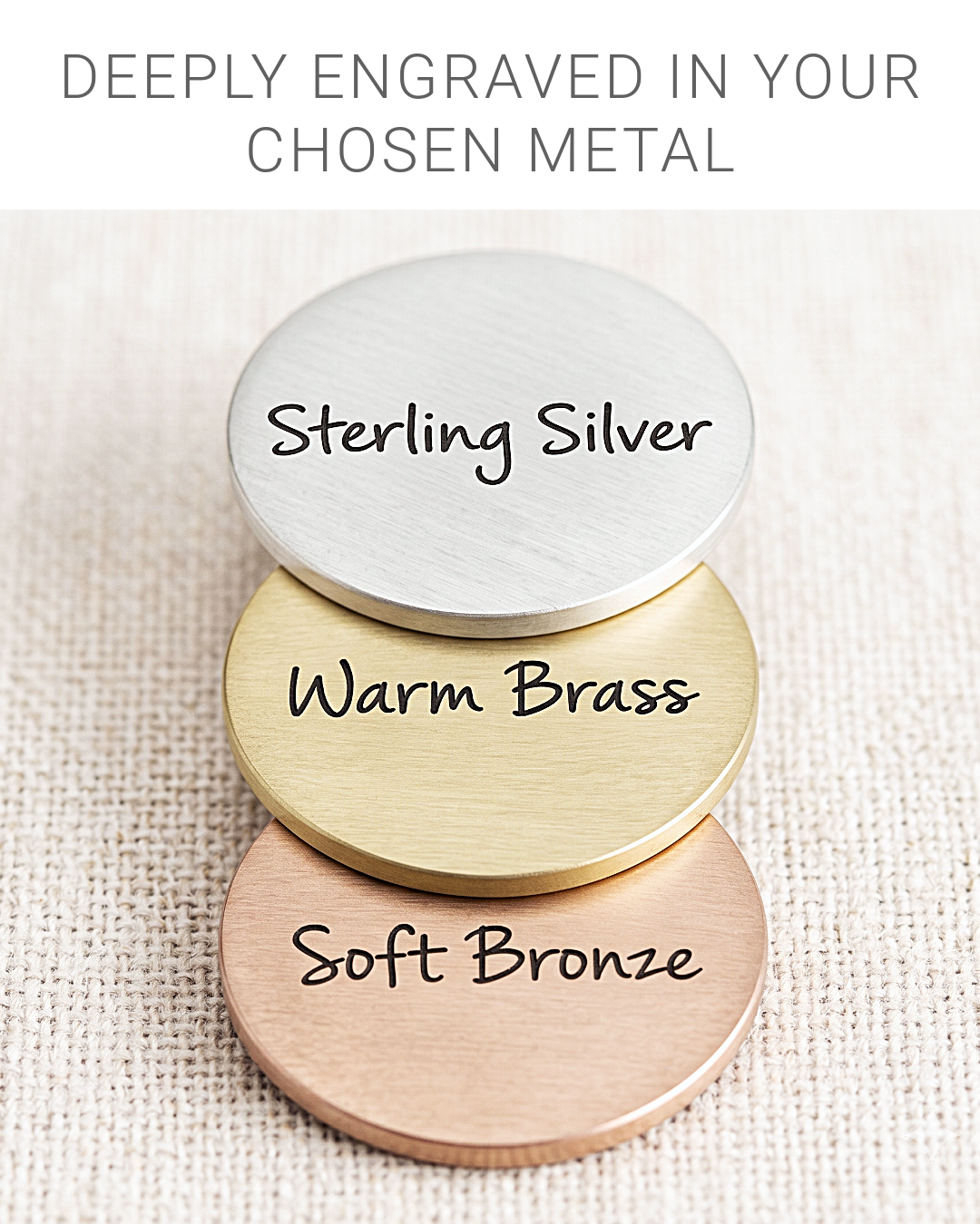 Three round engraved metal nameplates in Sterling Silver, Warm Brass, and Soft Bronze stacked on a textured beige fabric background, with text at the top reading 'DEEPLY ENGRAVED IN YOUR CHOSEN METAL'.