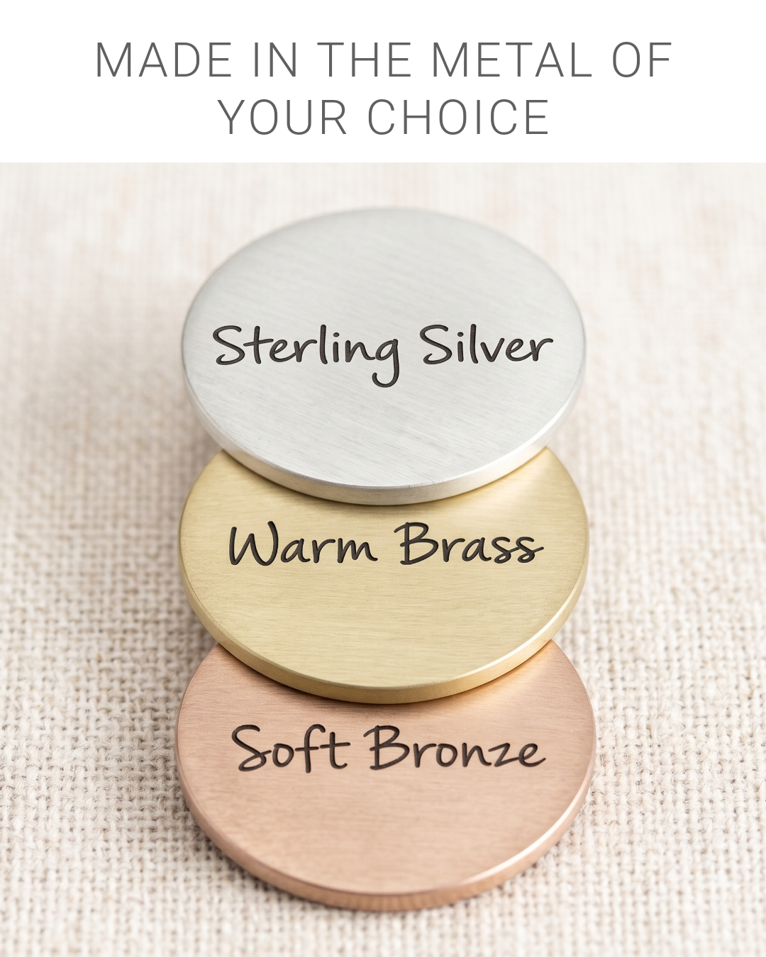 Three round metal nameplates in Sterling Silver, Warm Brass, and Soft Bronze stacked on a textured beige fabric background, with text at the top reading 'MADE IN THE METAL OF YOUR CHOICE'.