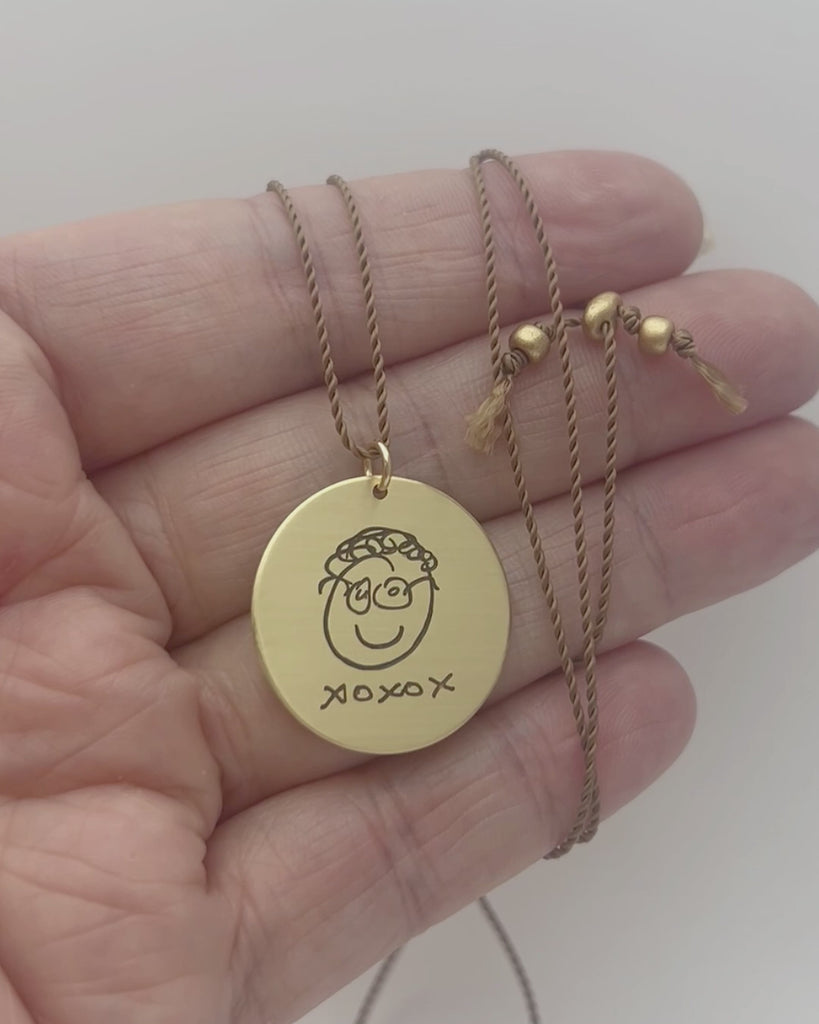Large round, gold pendant engraved with handwriting and a doodled character. Modeled and moving. 