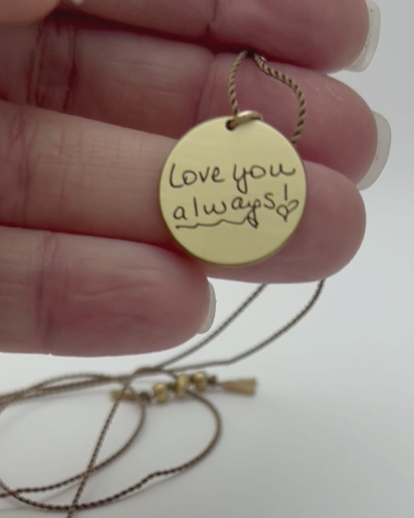 Round brass Leah pendant engraved with handwriting and held in hand. 